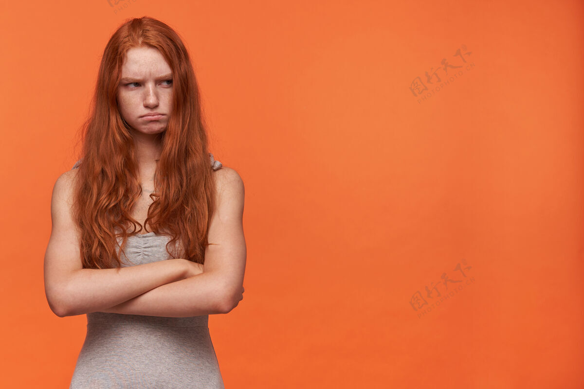 20多岁一张室内照片 年轻的红发可爱的女人一边看一边不高兴地皱着眉头 穿着休闲服站在橙色背景上 双臂交叉手臂长女士