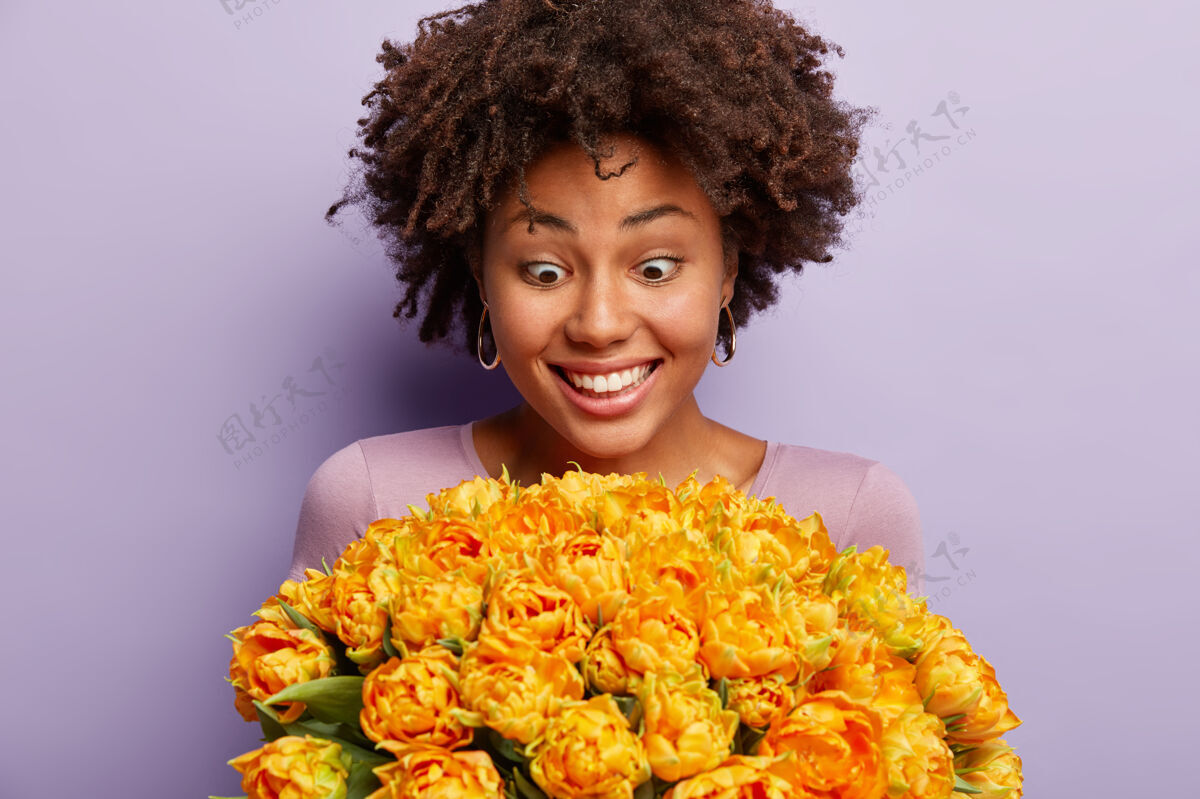 个性特写镜头中的快乐惊喜的黑皮肤年轻女子盯着一大束鲜花 不敢相信这礼物是送给她的 隔着紫色的墙哇 多漂亮的郁金香啊女人花难以置信