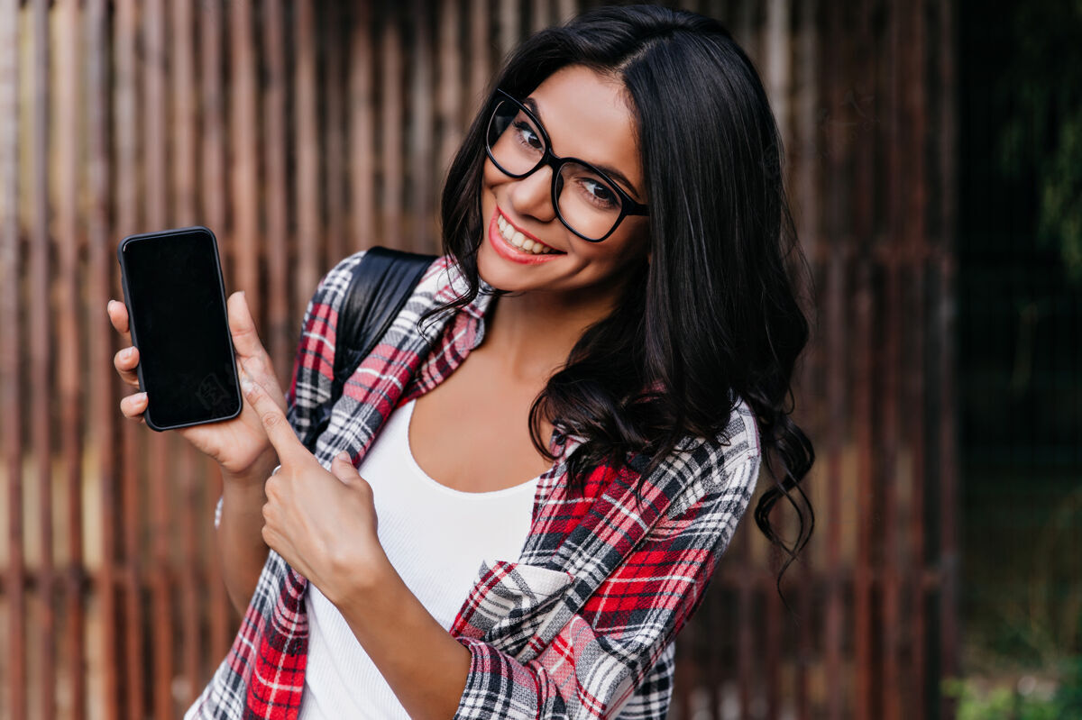 年轻漂亮的卷发拉丁女人展示新手机戴着眼镜的可爱黑发女孩的户外肖像 拿着智能手机在木墙上摆姿势休闲小玩意快乐