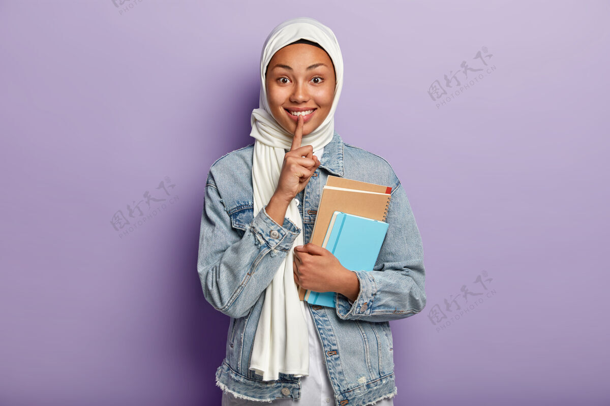 伊斯兰教快乐的穆斯林妇女的上身镜头 表情愉快 做嘘手势 露出白色的牙齿 拿着螺旋形的记事本 戴着白色的围巾 牛仔夹克 隔着紫色的墙记事本青少年无声