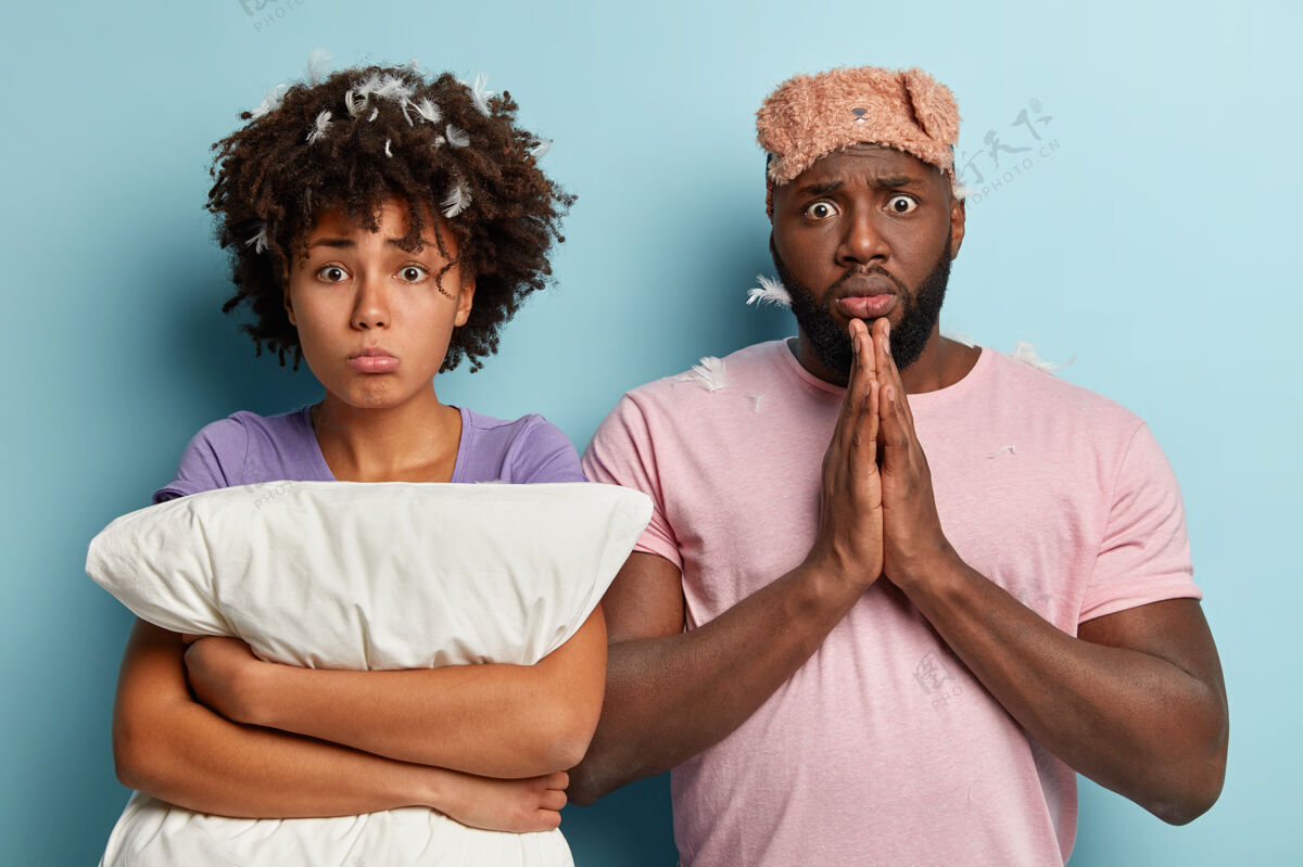 剃胡子沮丧的黑人女人留着非洲发型 抱着白色的枕头 震惊的黑人男人双手合十 眼睛有窃听器 戴着睡眠面罩 站得很近睡眠和休息的概念不安惊喜昏迷