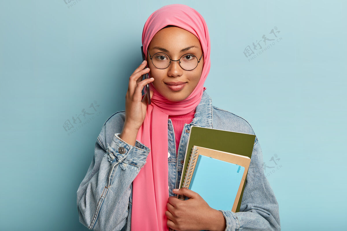 阿拉伯语人 科技 种族 沟通观念戴着穆斯林传统头巾的漂亮女孩与团友通电话 讨论未来项目 拿着两个螺旋笔记本 在室内摆姿势围巾高兴伊斯兰