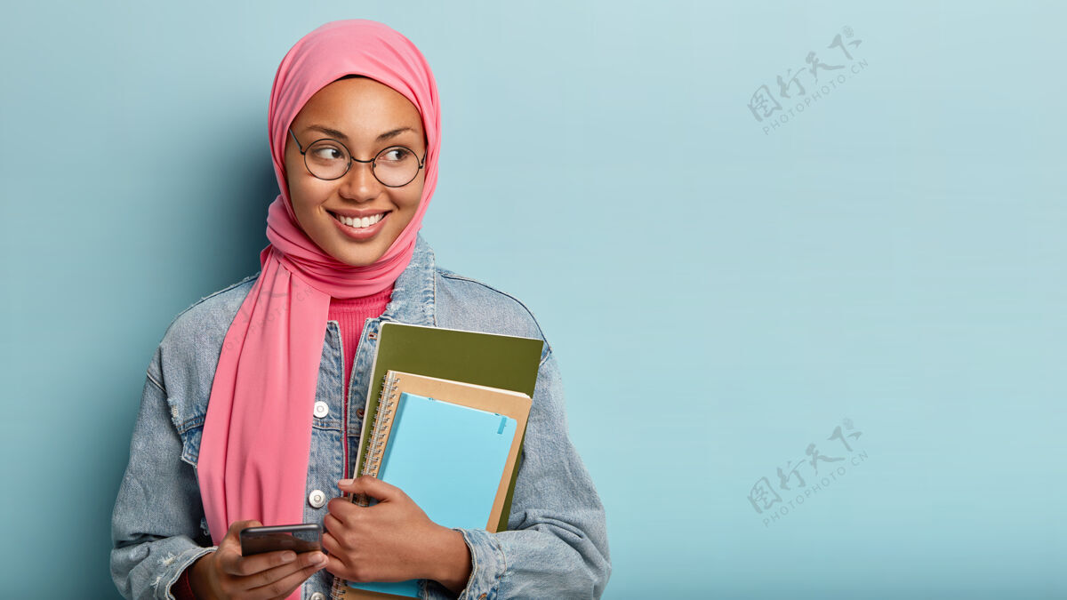 宗教快乐的穆斯林学生在手机上打短信 拿着记事本 带着快乐的表情专注在一边 穿着牛仔夹克 隔着蓝色的墙壁 读着有趣的文章短信满意智能手机