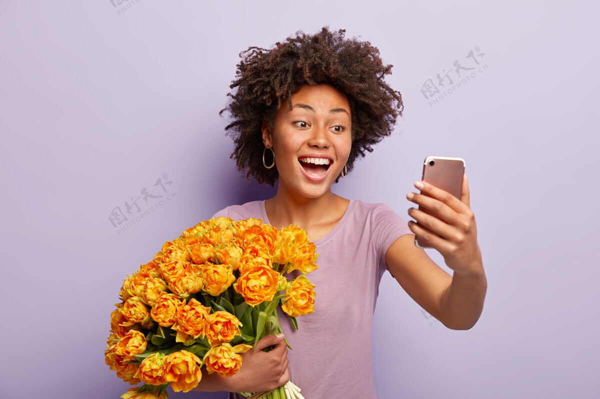 郁金香快乐的黑皮肤女士表达真挚的感情和情感 在社交网络上自拍分享照片 捧着漂亮的大束橙花 穿着休闲t恤 手机快乐高兴