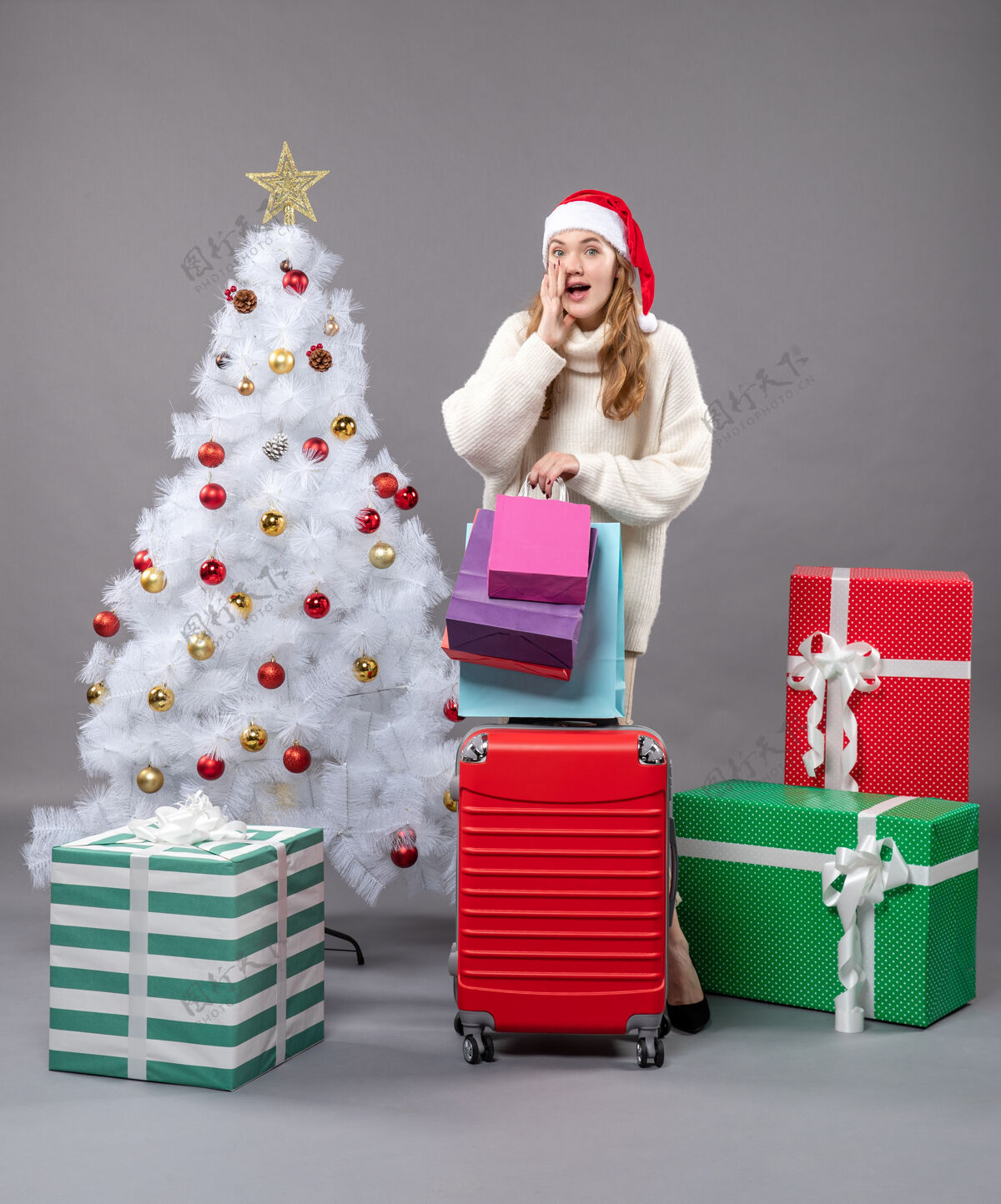 灰色正面图惊讶的金发女孩戴着圣诞帽 手里拿着红色的手提包和购物袋圣诞帽圣诞树盒子