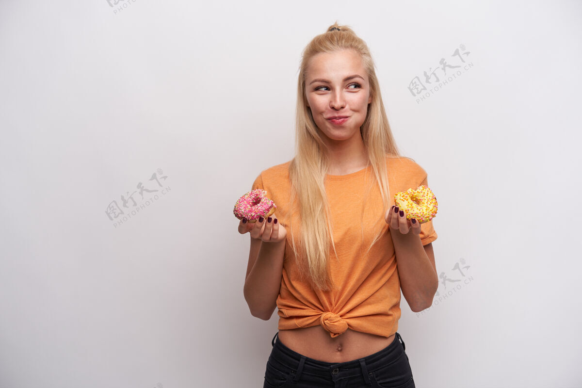饮食在室内拍摄的年轻快乐的长发金发女士一边嚼着被咬的甜甜圈一边积极地看着一边 举起手拿着捐款 一边站在白色的背景上手摆姿势女性