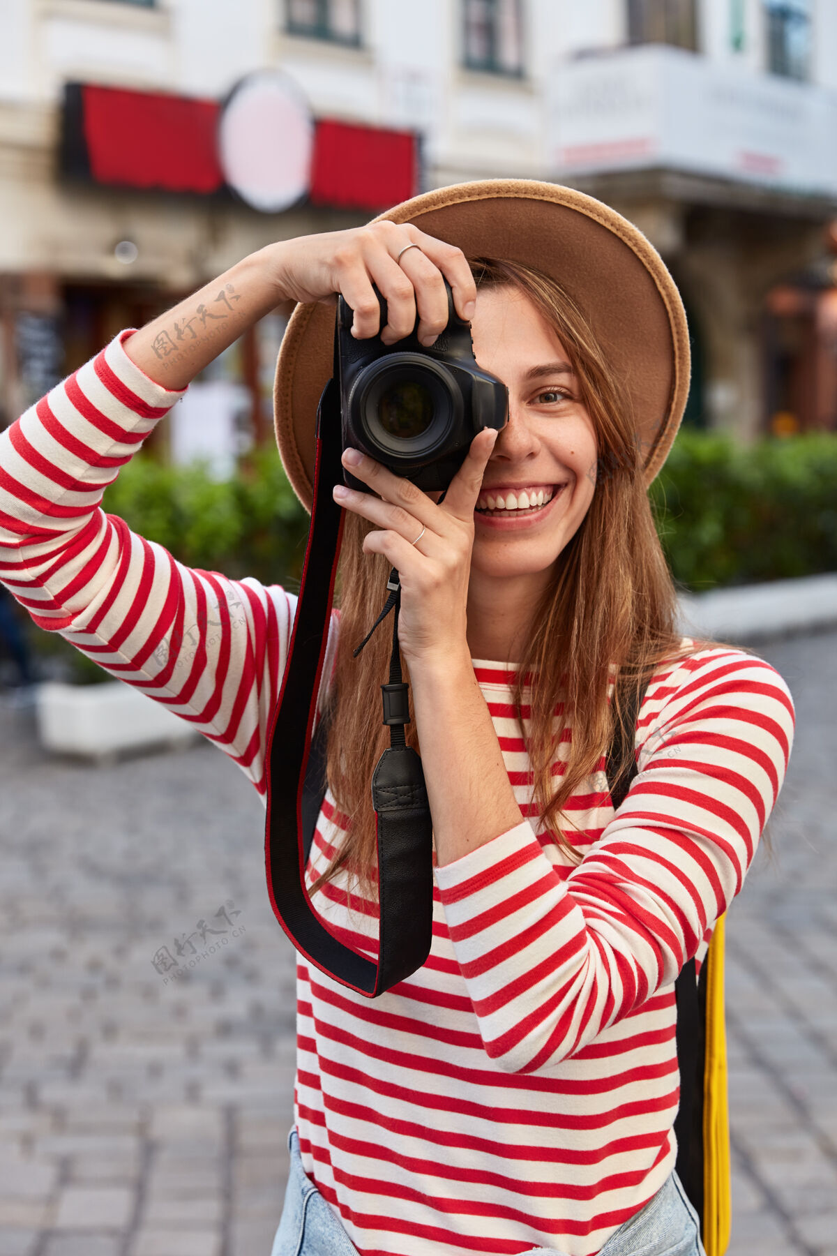 广场高兴的游客在相机上专业拍照 笑容灿烂 关注美丽的地标 漫步在市中心 戴着帽子城镇条纹帽子