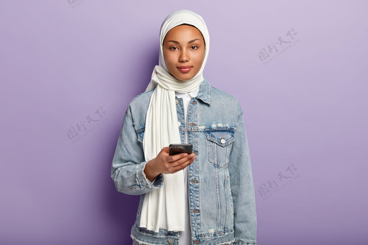 伊斯兰教一张美丽的阿拉伯女人的照片 皮肤黝黑健康 在现代手机上发短信 在帖子下面读评论 戴着白色头巾 穿着牛仔外套 模特们站在紫色的墙上穆斯林宗教肖像阿拉伯语智能