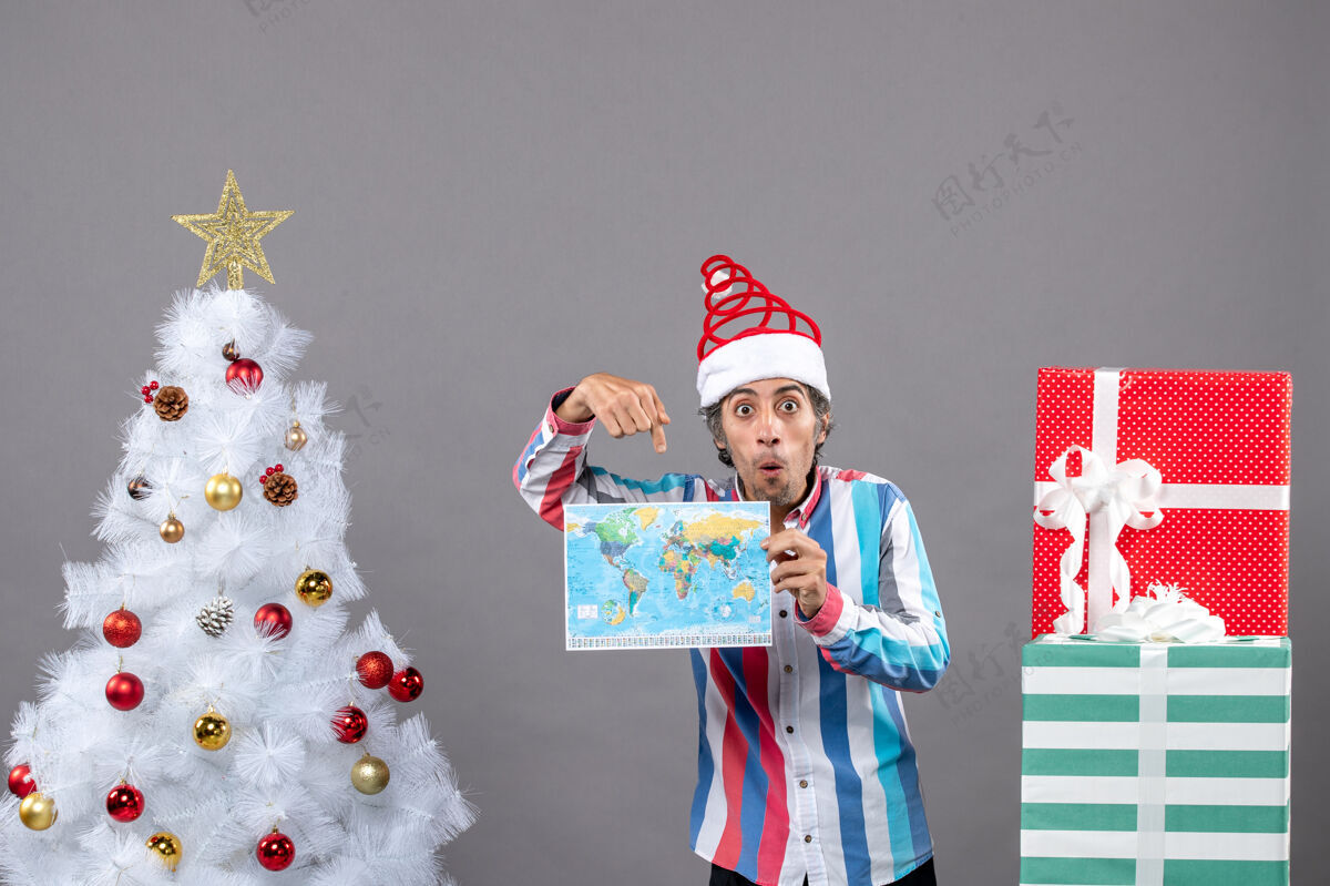 惊讶的男人前视图惊讶的男子螺旋弹簧圣诞帽和条纹衬衫显示地图附近的白色圣诞树和复制空间人家伙圣诞老人