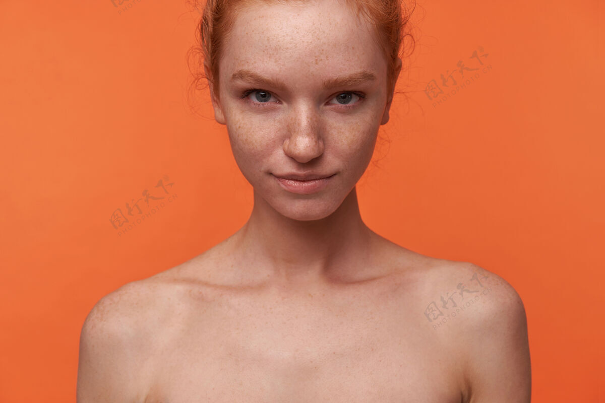 手摄影棚拍摄的红头发打结的美丽小姐站在橙色背景上 双手放在地上 扬起眉毛对镜头轻轻微笑人类情感面部表情肢体语言语言冷静折叠