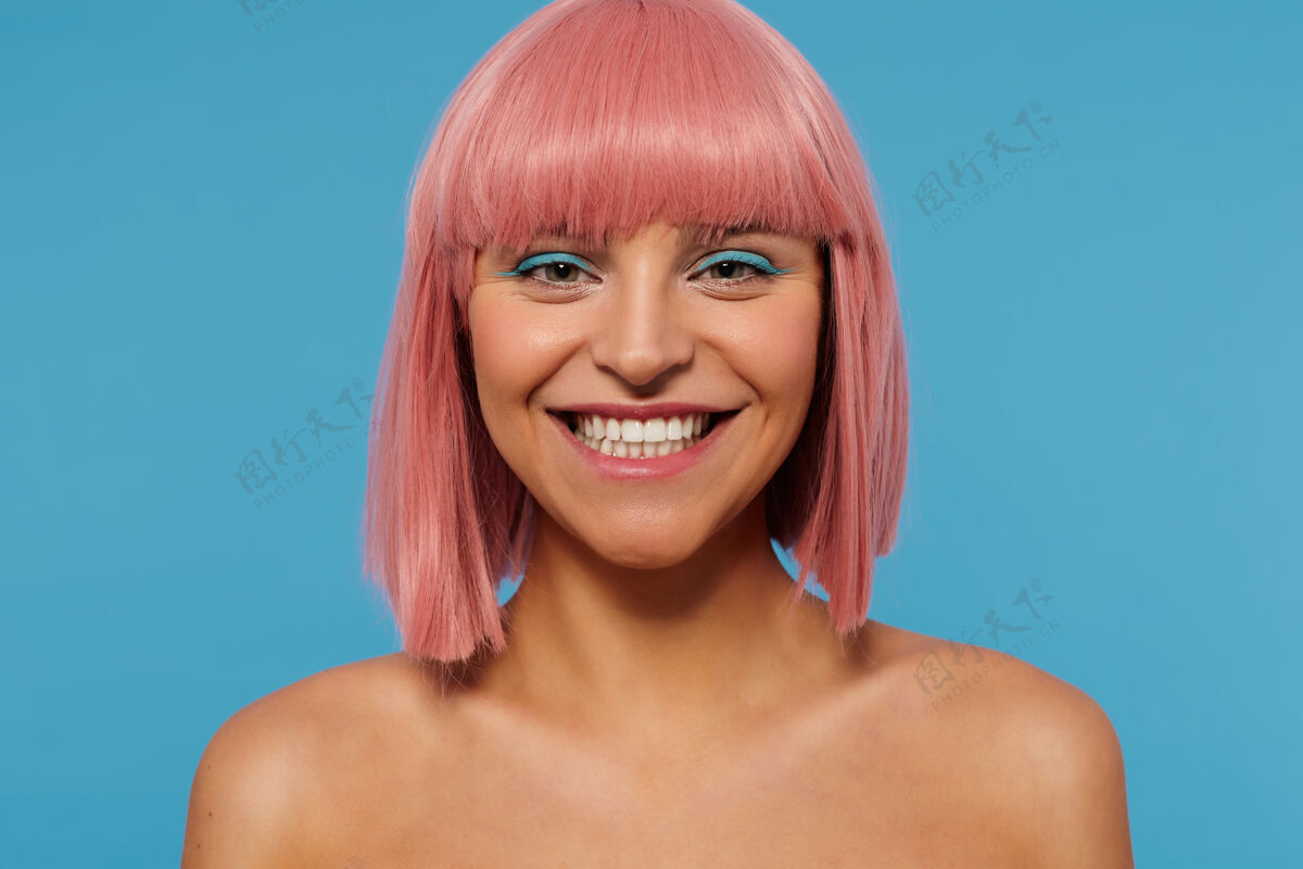 鲍伯特写镜头中的快乐年轻迷人的粉红色头发的女性与彩妆显示她完美的白色牙齿 同时愉快地看着相机 在蓝色的背景隔离箭头表情肩膀