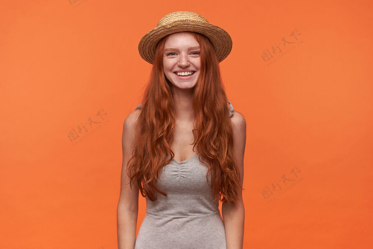 肖像身着灰色衬衫 头戴夏季草帽的迷人的长发狐狸精女郎站在橙色背景下 双手放在地上 积极地看着镜头女性衬衫表情