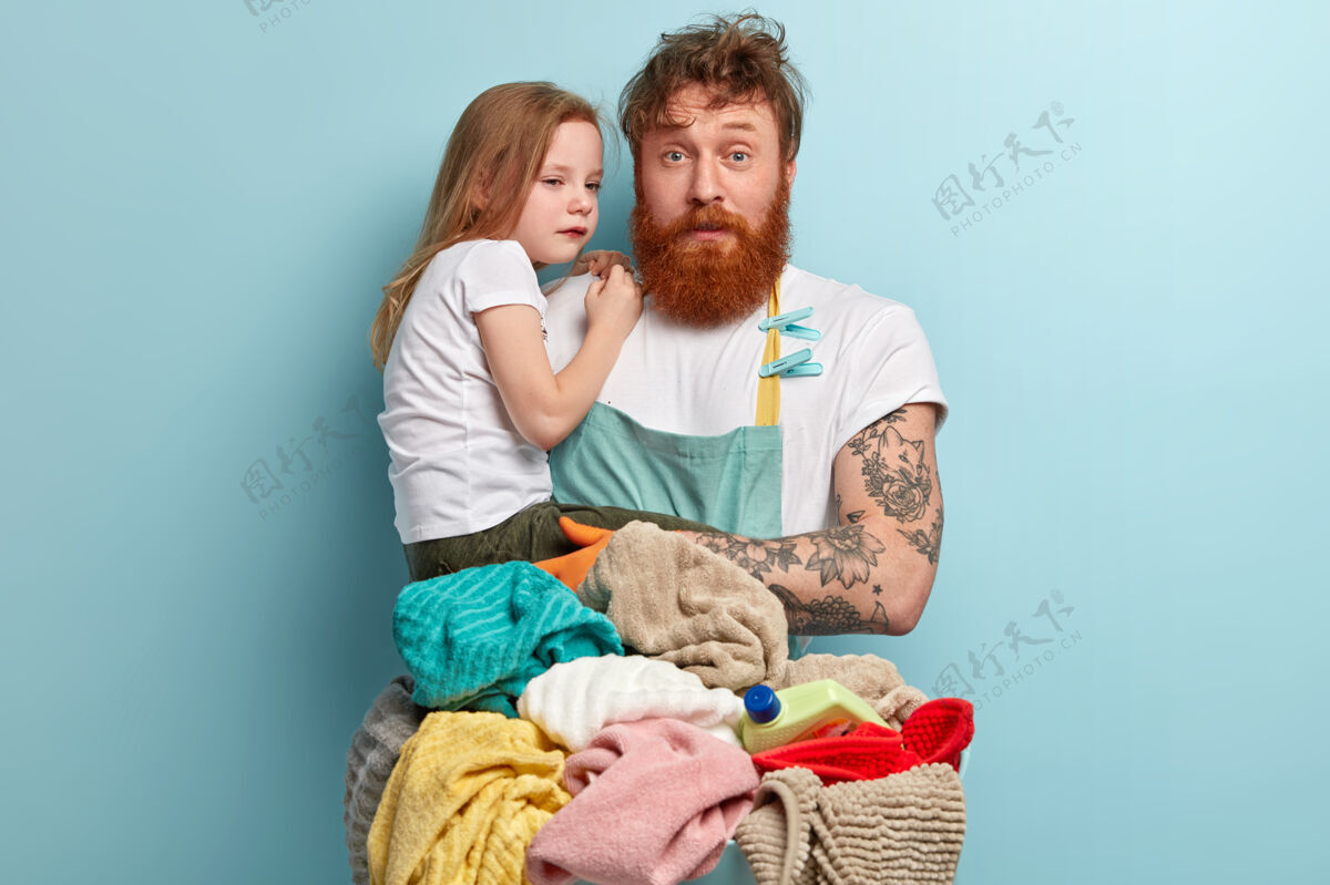 红发忙碌狡猾的单身爸爸试图逗弄哭闹的孩子 神情迷惑不解 穿着围裙 洗衣服 有很多家务活 隔着蓝色的墙父爱和商业理念父亲父母手套