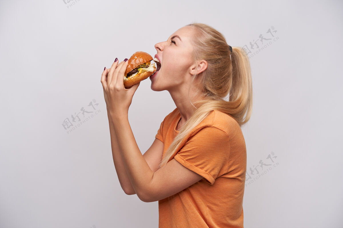 饮食饥饿的年轻长发金发女郎的后视图 随意的发型 贪婪地吃着新鲜的大汉堡 站在白色的背景下 闭着眼睛垃圾室内汉堡