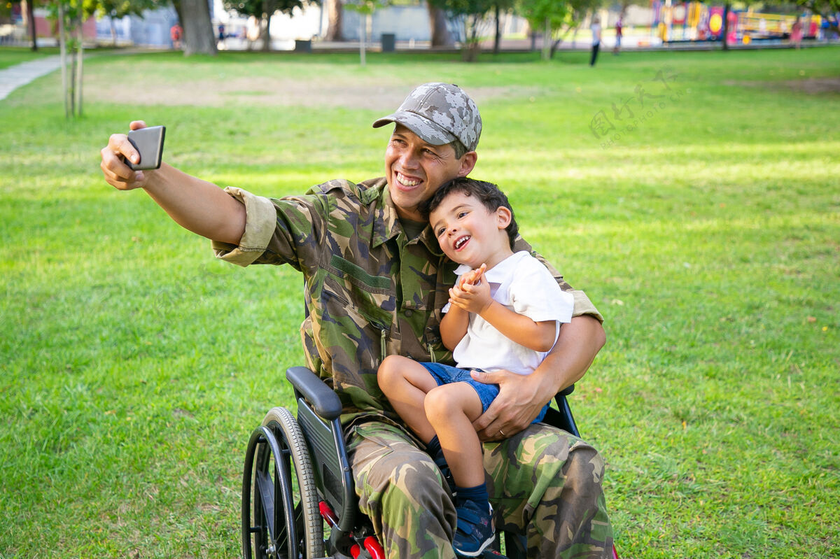父亲快乐的残疾军人爸爸和他的小儿子一起在公园里自拍男孩坐在爸爸的膝盖上退伍军人或残疾概念父母椅子伪装