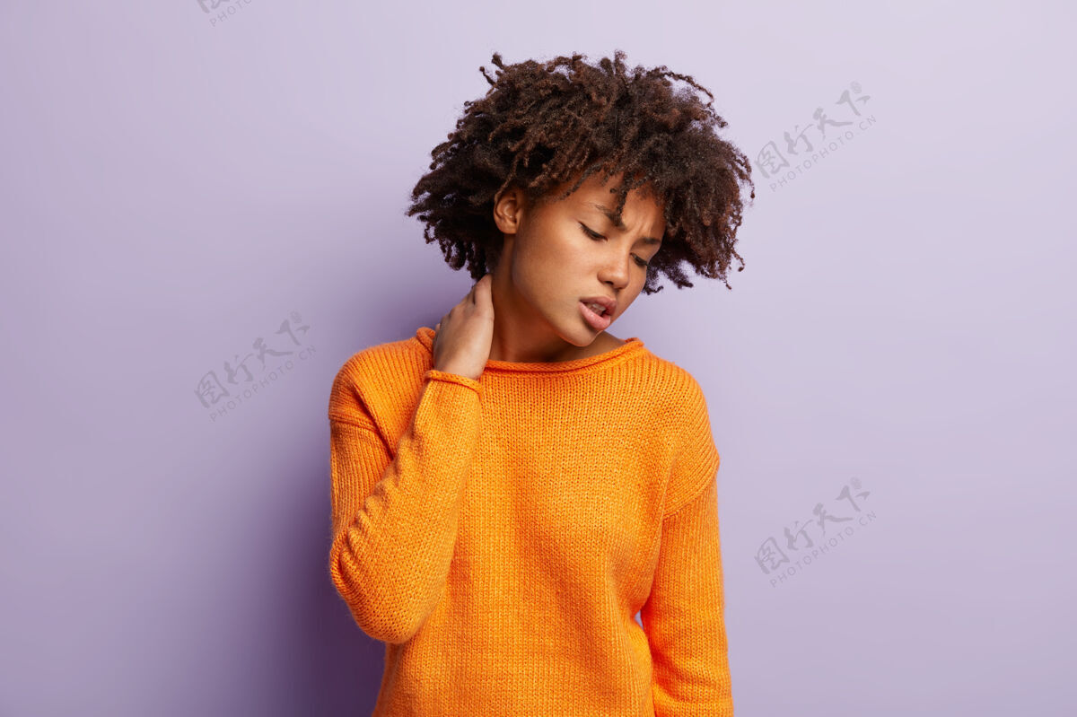 种族不满的卷发女人的照片手放在脖子上 忍受着可怕的疼痛 工作努力 有久坐的生活方式 浓密的卷发 穿着橙色的套头衫 模特在紫色的墙上室内过度劳累不满