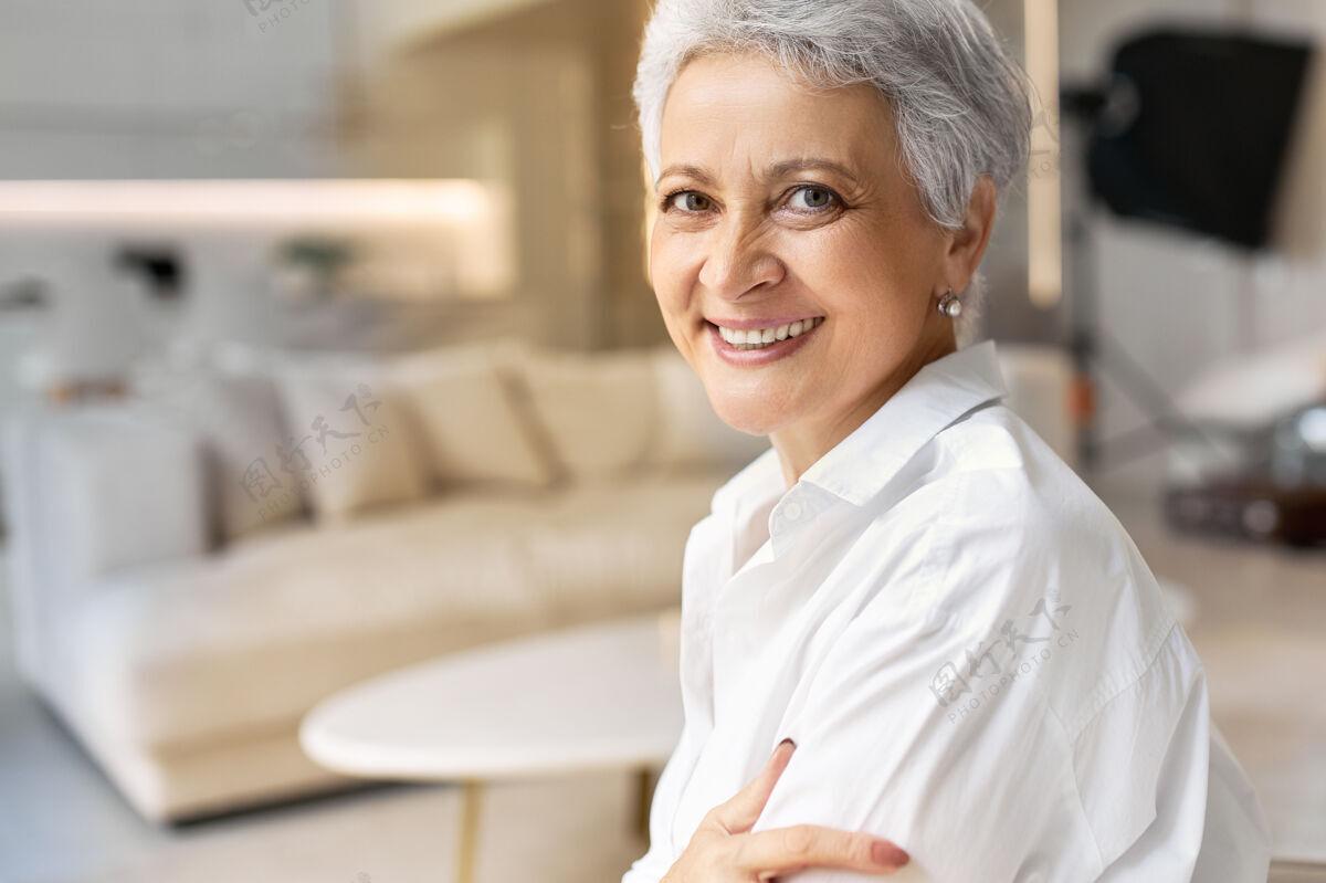 工作50岁的退休老太太 满脸雀斑 头发灰白 穿着白衬衫 面带笑容 在时尚的室内背景下摆姿势现代自信室内
