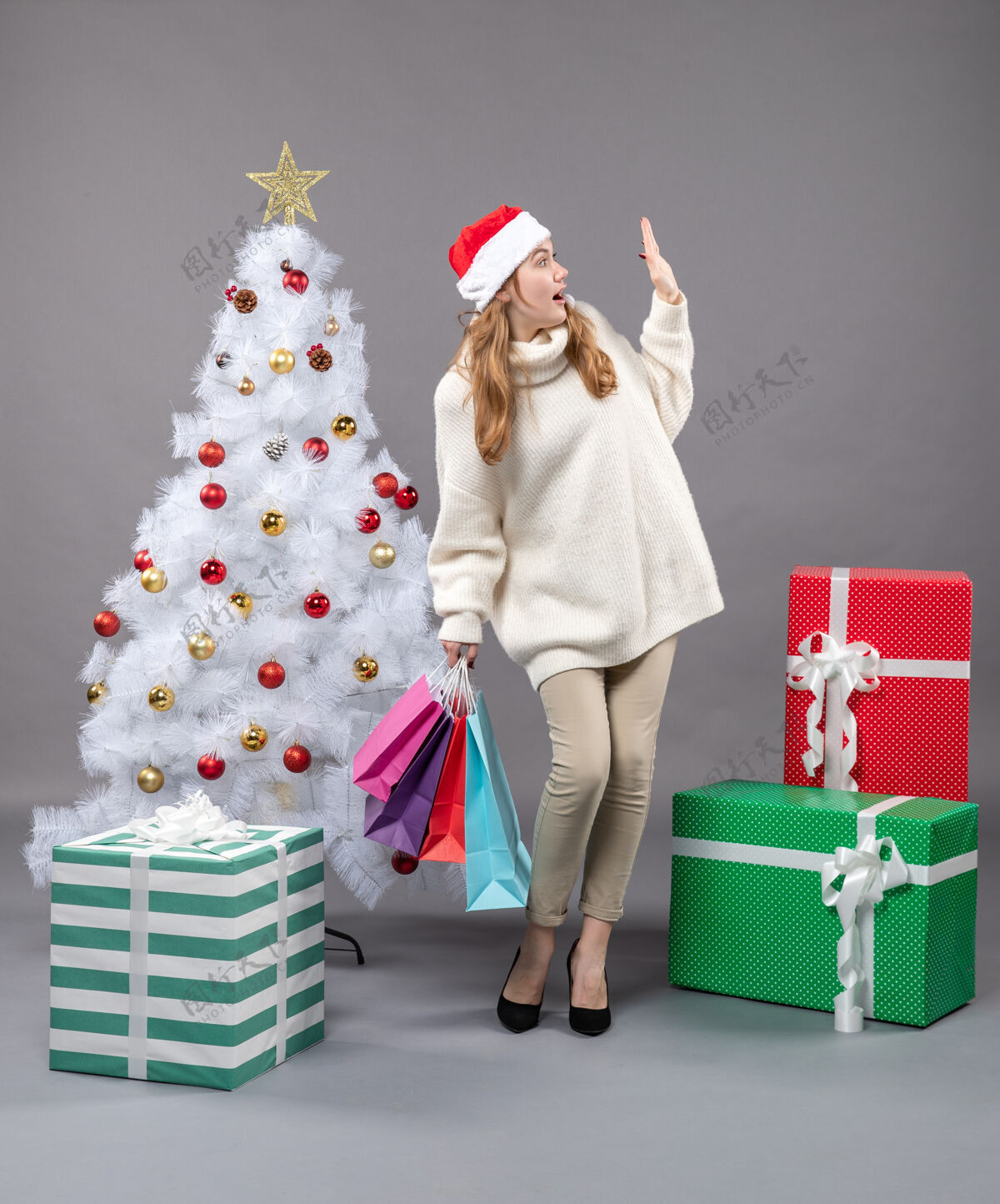 礼物正面图令人惊讶的圣诞妇女戴着圣诞帽 手里拿着许多五颜六色的购物袋商店灰色五颜六色