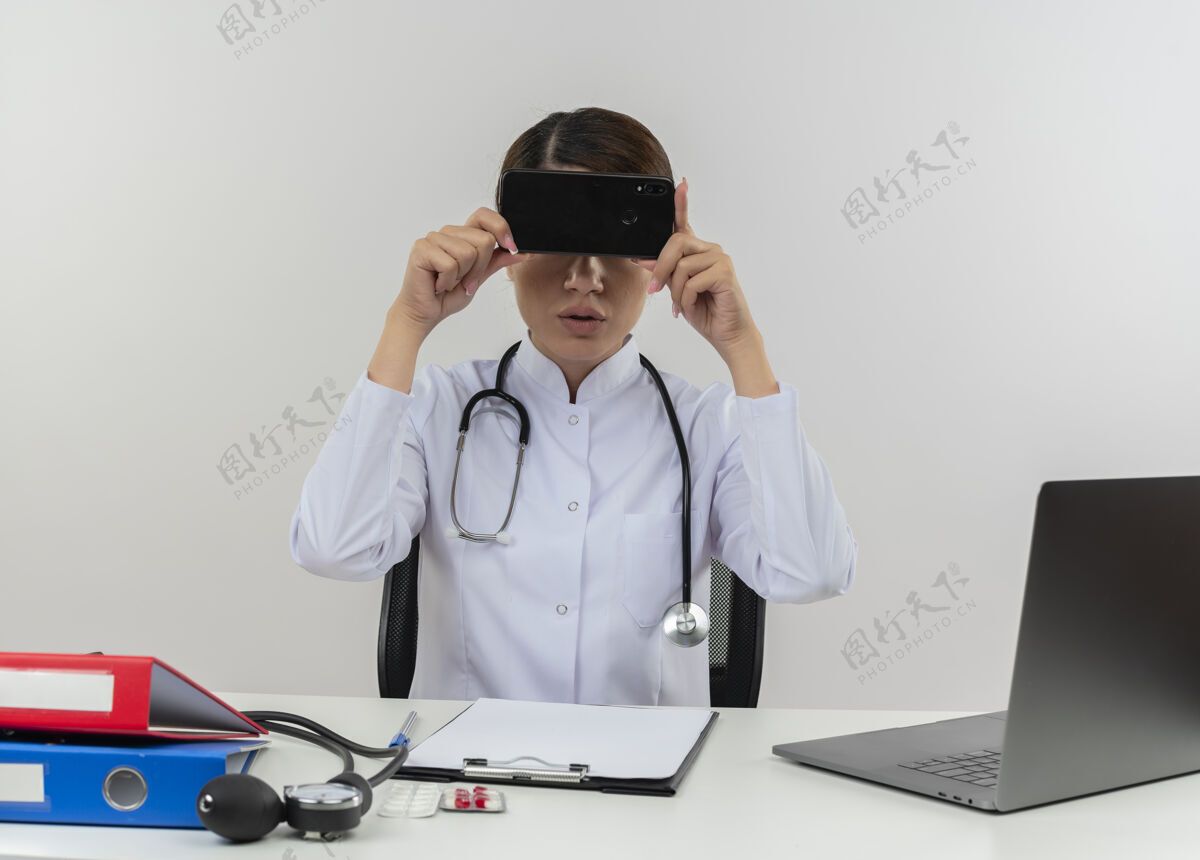电脑年轻的女医生穿着带听诊器的医用长袍坐在办公桌旁用电脑和医疗工具捂着眼睛用手机复印医生办公桌坐着