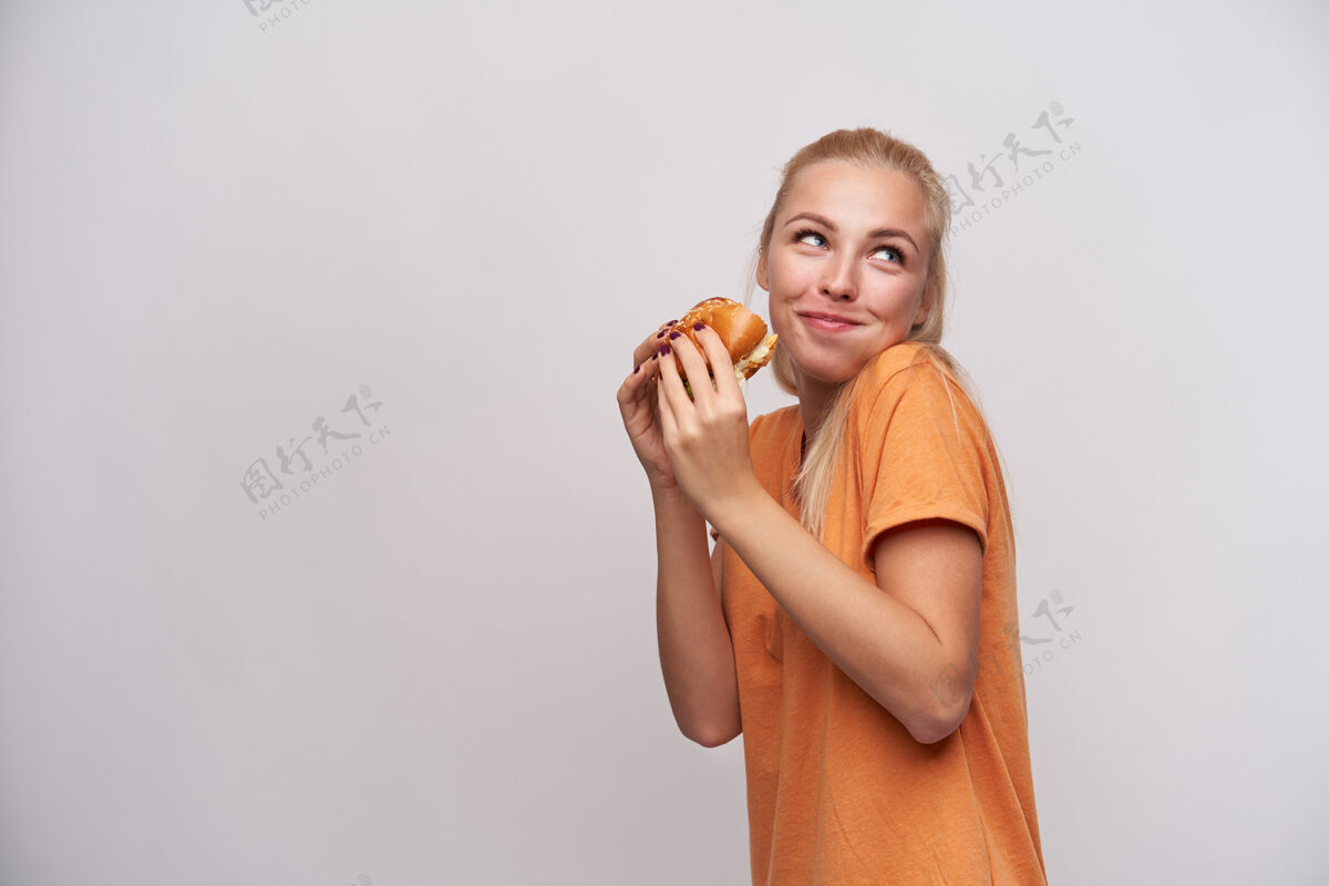 快餐摄影棚拍摄的饥饿高兴的年轻金发女性穿着橙色t恤正面看着一边 嘴里塞满了食物 拿着新鲜的汉堡 站在白色背景上食物情绪表情