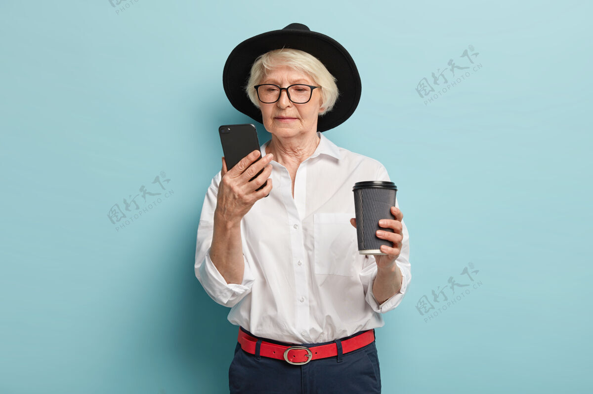 养老金严肃的老年妇女专注于智能手机设备 在互联网上搜索信息 在线阅读文章 喝咖啡去 退休人员使用简单的应用程序 穿着时尚的服装 在室内摆姿势风格短信腰带