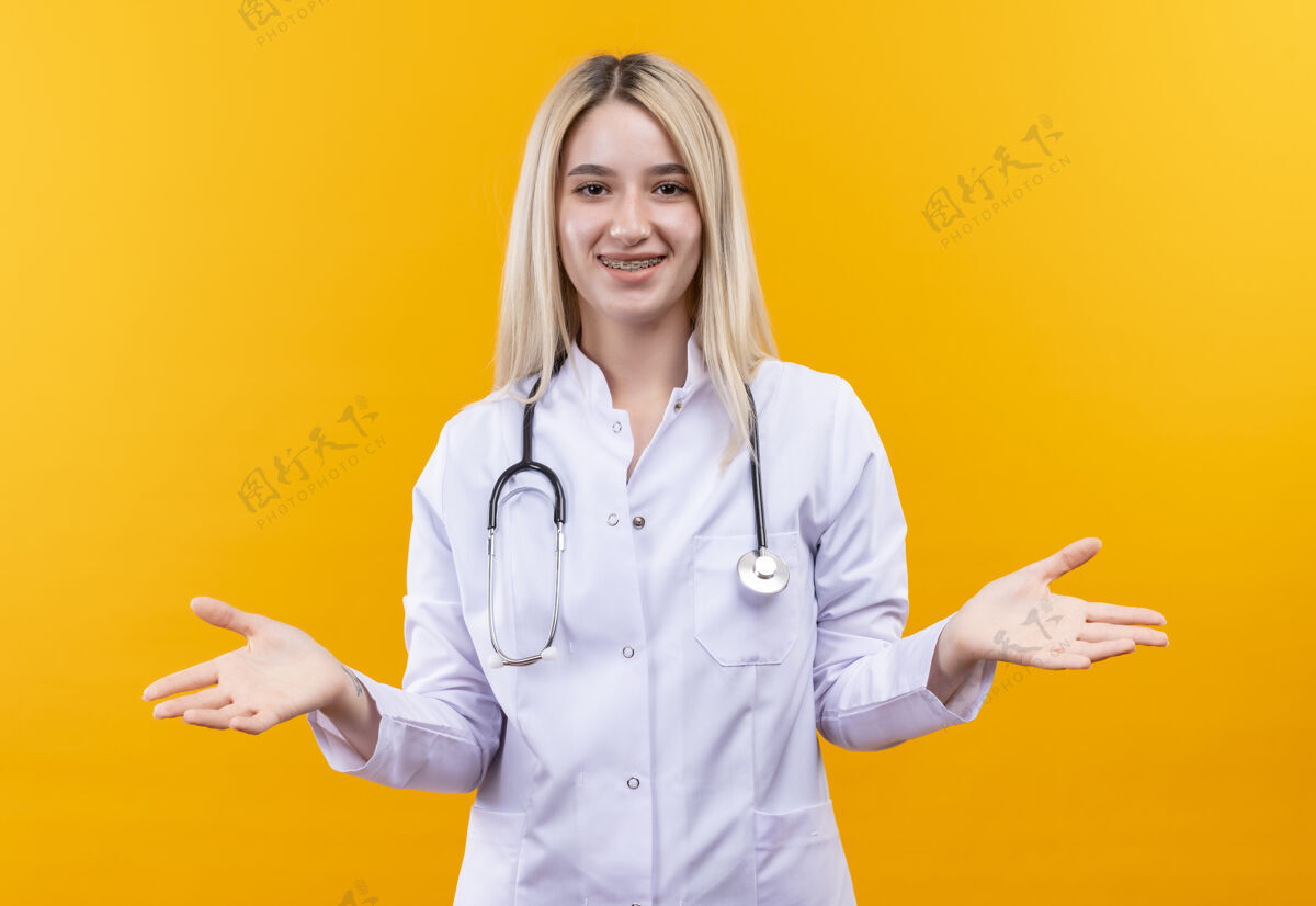 手微笑的医生穿着医用长袍戴着牙套戴着听诊器的年轻女孩在孤立的黄色背景上展开双手牙科黄色传播