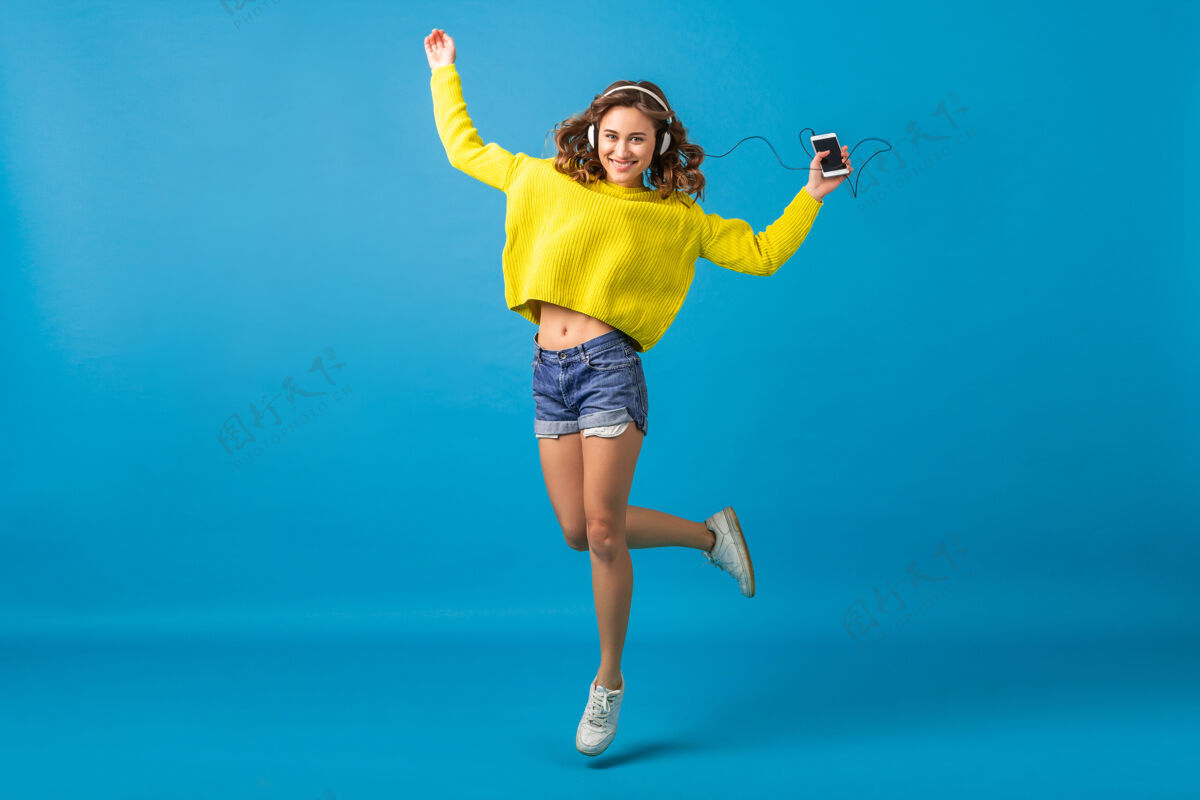耳机迷人的笑容可掬的快乐女人戴着耳机跳着舞听着音乐 穿着时髦的服装 隔离在蓝色的工作室背景下 穿着短裤和黄色的毛衣蓝色五颜六色长度