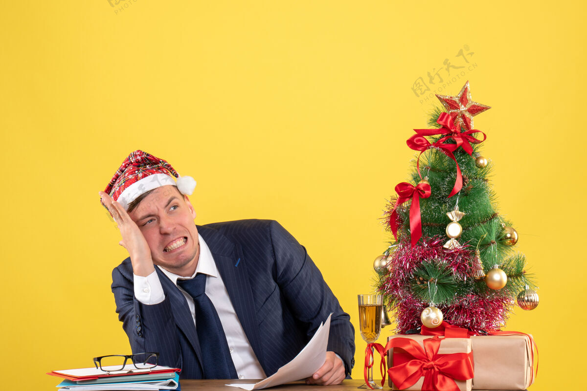 人们一个戴着圣诞帽的忧心忡忡的男人坐在圣诞树旁的桌子旁 拿着黄色的礼物西装圣诞节男性