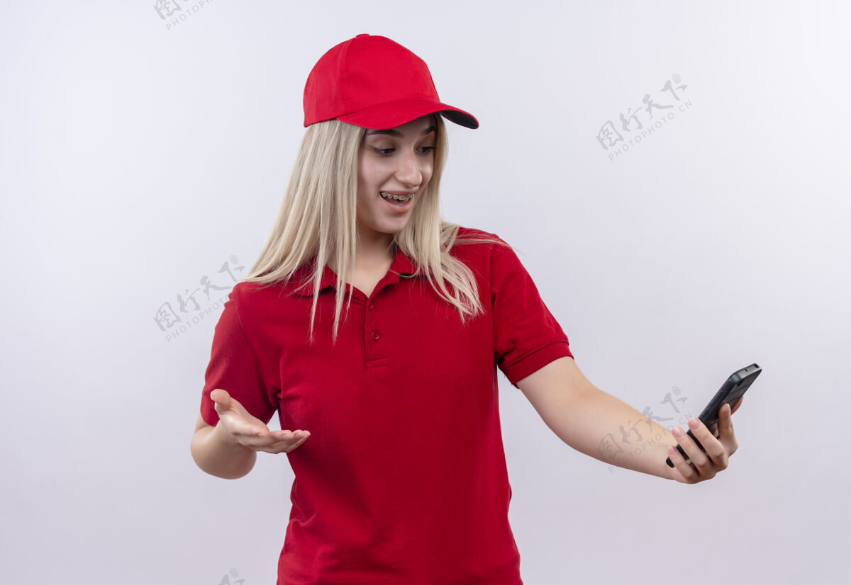 送货一个穿着红色t恤和帽子的年轻女孩在孤立的白色背景下看着手上的手机手惊讶穿
