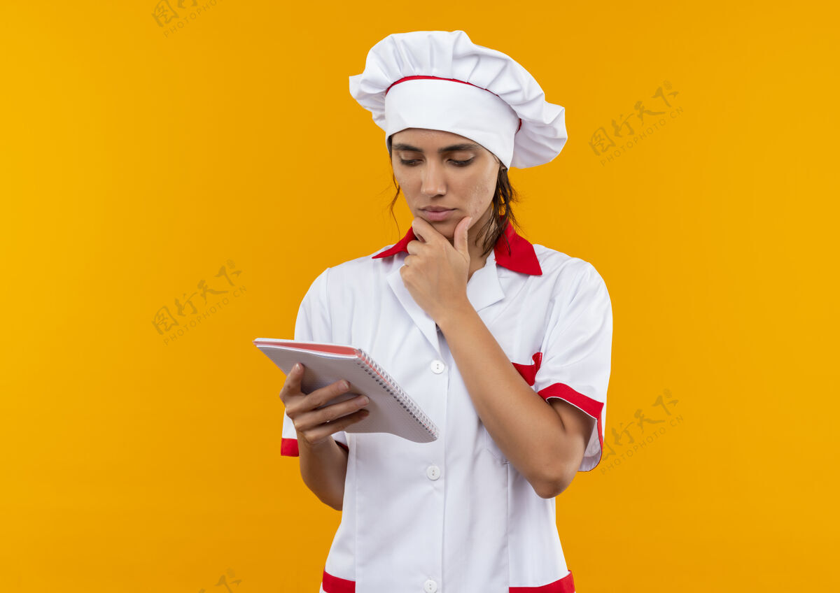拿想着年轻的女厨师穿着厨师制服拿着笔记本看着 手放在下巴下面留着复印空间手厨师下巴