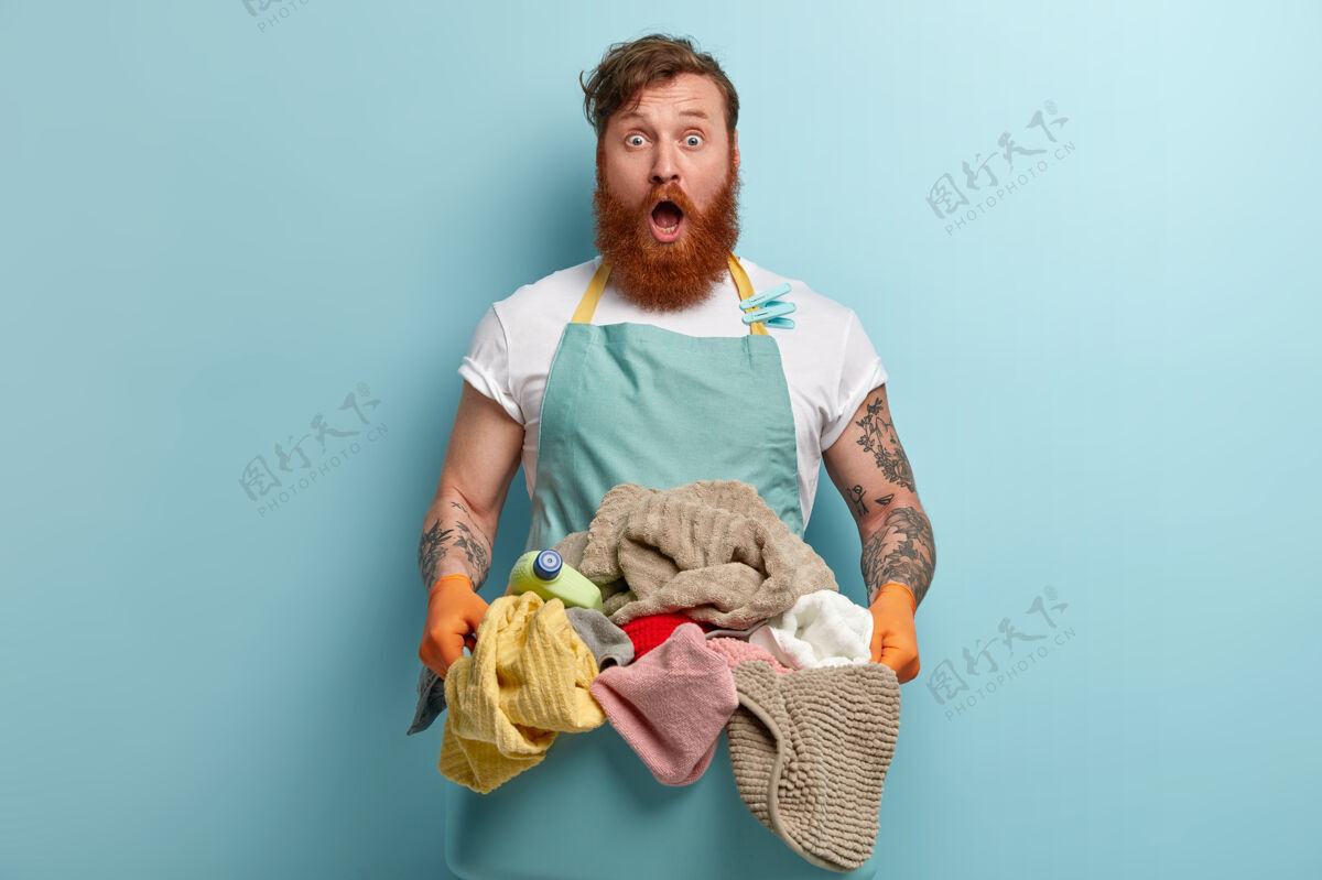 红发惊惶失措的红发大胡子男人穿着休闲的蓝色围裙 手里拿着一盆展开的衣服情绪化洗衣家伙