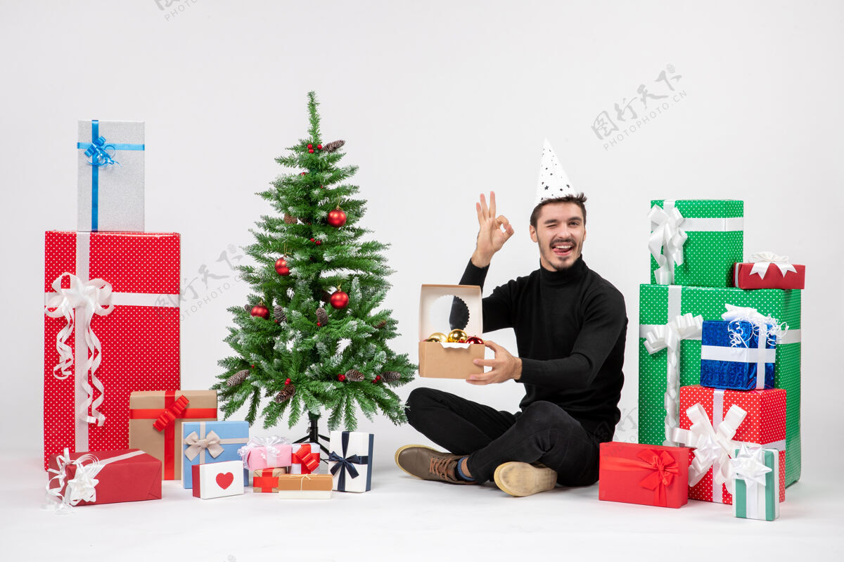 节日正面图：年轻人围坐在节日礼物旁边 手里拿着白色墙上的玩具包快乐人圣诞树