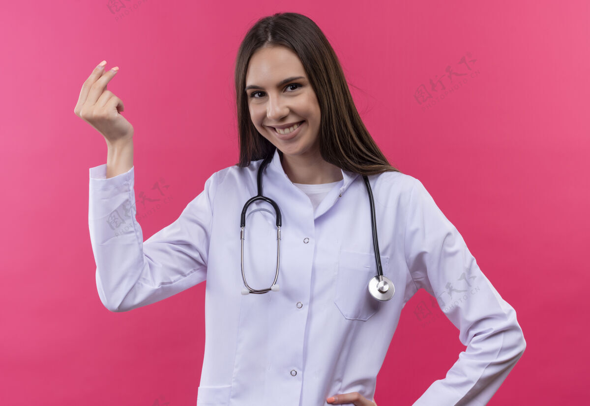礼服微笑的年轻医生女孩穿着听诊器医用长袍 在孤立的粉红色背景上显示提示手势听诊器医生粉红