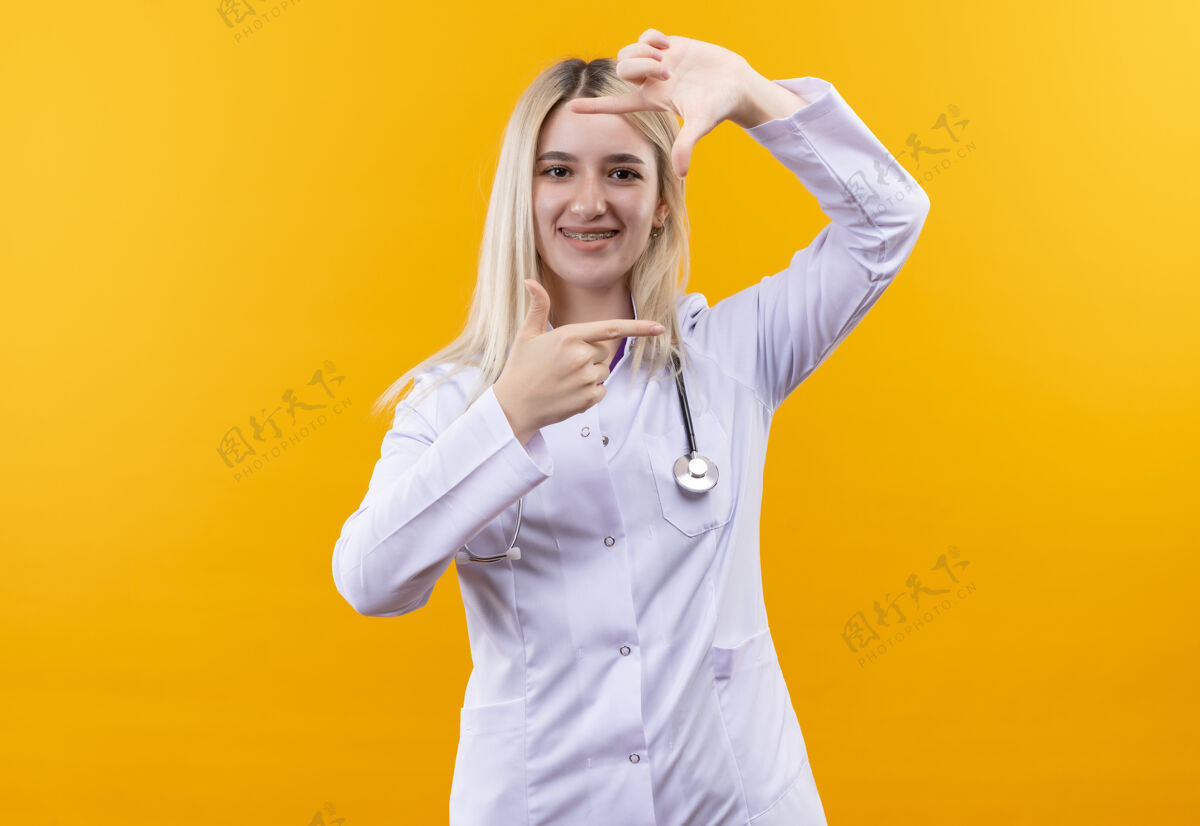女孩微笑的医生穿着医用长袍戴着听诊器和牙套的年轻女孩在孤立的黄色背景上展示着照片的姿势手势听诊器穿着