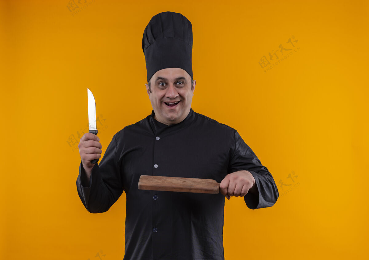 厨师惊讶的中年男厨师穿着厨师制服拿着刀和砧板在黄色背景上复制空间中年厨师切割