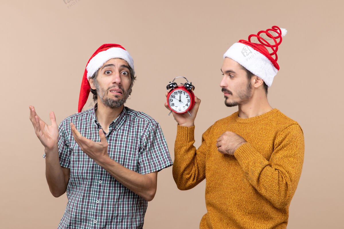 手持前视图两个男人一个拿着闹钟 另一个在米色孤立的背景上混淆它时钟闹钟前面