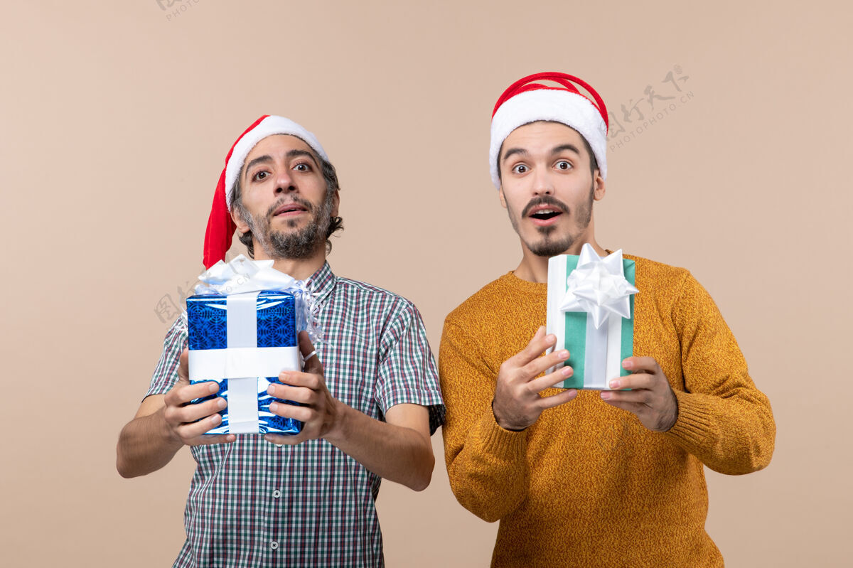 微笑正面图两个满意的家伙戴着圣诞帽 拿着圣诞礼物 背景是米色的团体礼物两个