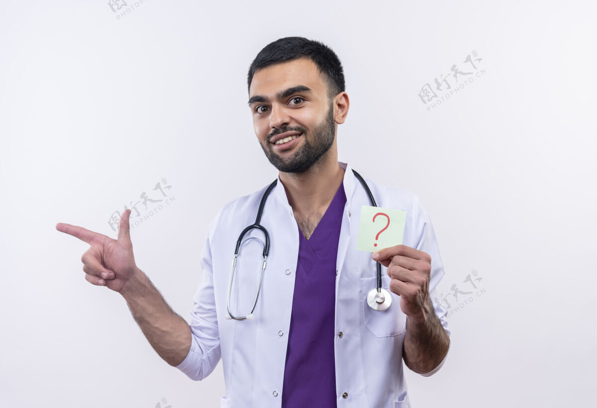 年轻微笑着的年轻男医生穿着听诊器医用长袍拿着纸问号指着隔离的白色背景上的一边医生抱着问号