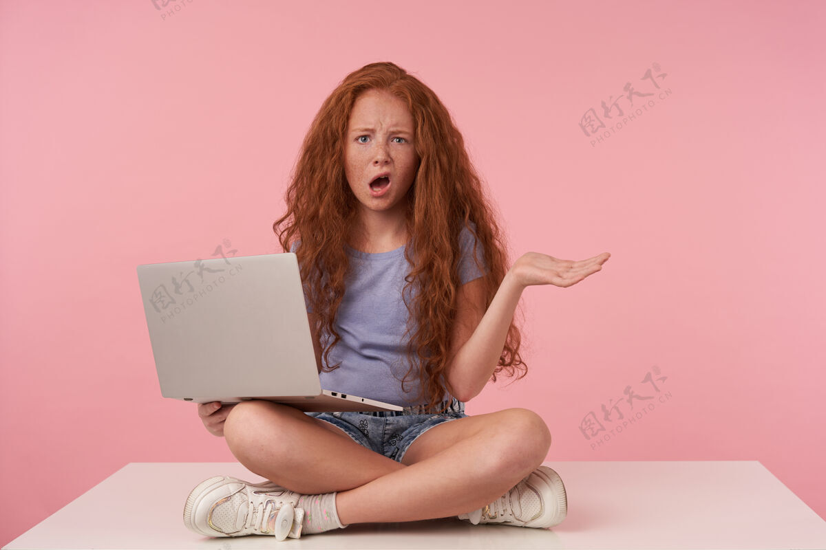 童年摄影棚拍摄的一个红发卷发女孩 一头长发 困惑地看着相机 腿上拿着笔记本电脑 撅着嘴抬起手掌 穿着蓝色t恤和牛仔裤短裤 在粉色背景上摆姿势漂亮孩子笔记本电脑