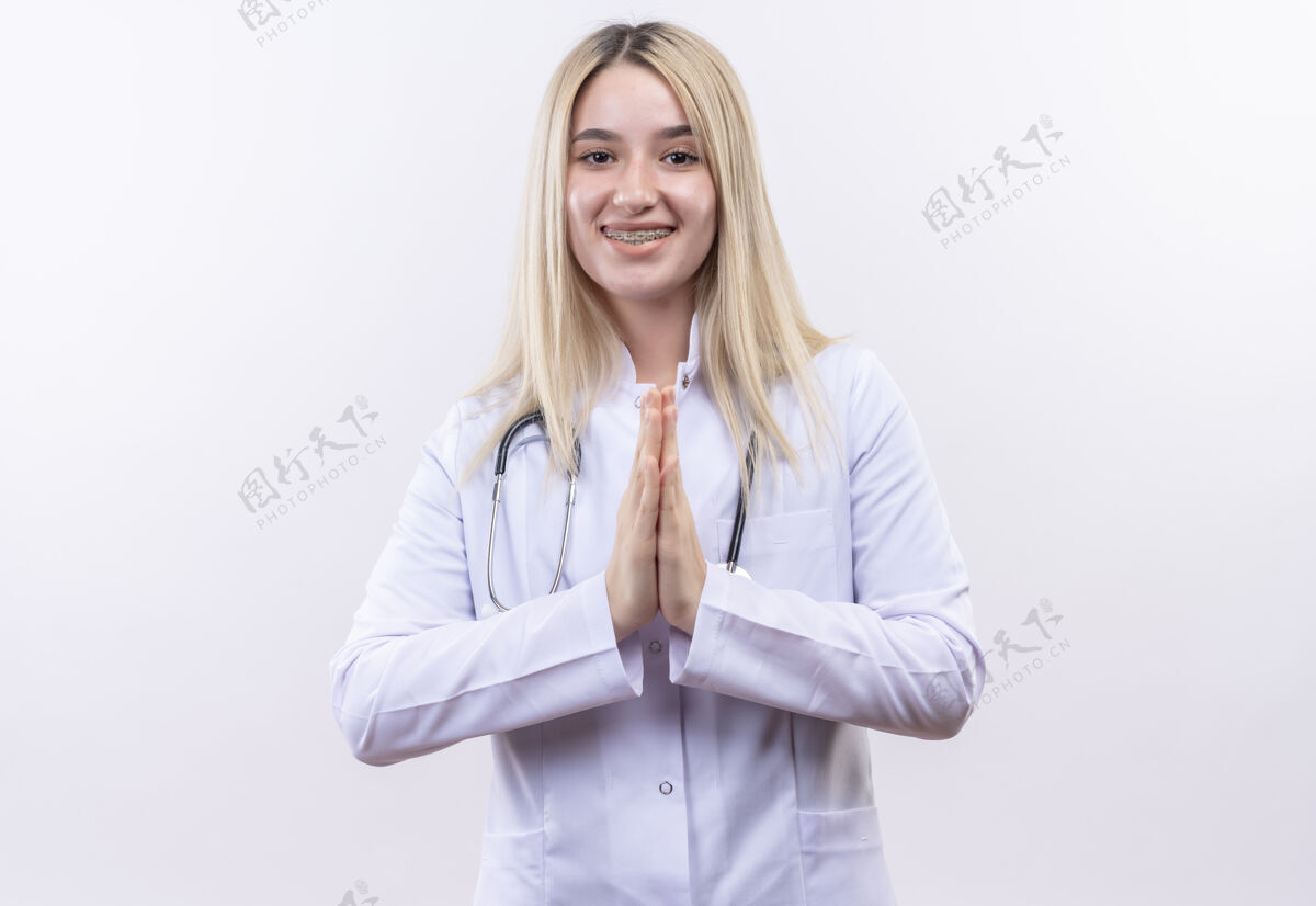 白人微笑的医生 年轻的金发女孩 戴着听诊器 穿着医用长袍 戴着牙套 在孤立的白色背景上做着祈祷的手势祈祷年轻穿着