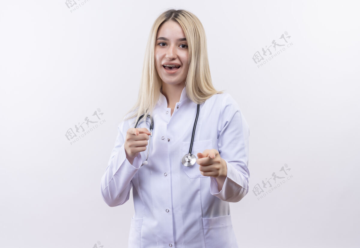 长袍微笑的医生 年轻的金发女孩 戴着听诊器 穿着医用长袍 戴着牙套 在孤立的白色背景上展示你双手的姿势牙科白色穿着