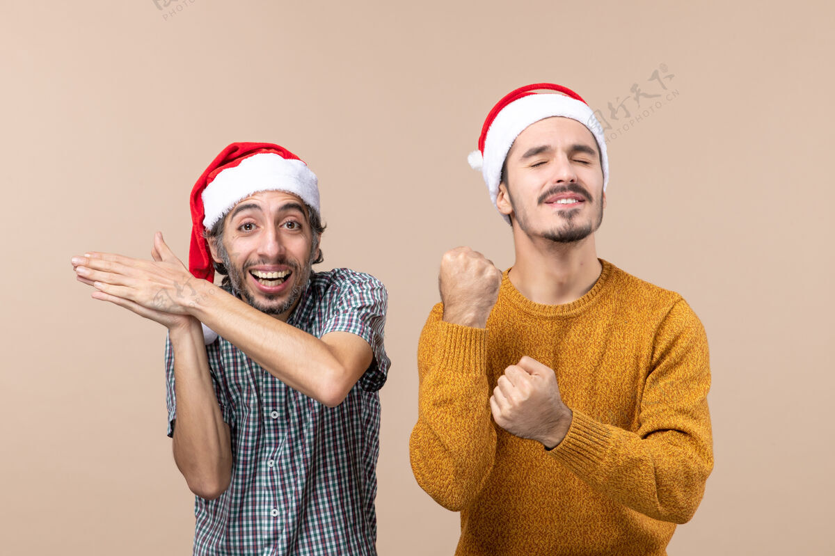 站着正面图两个戴着圣诞帽的家伙一个拍手 另一个闭着眼睛打拳站在米色的孤立背景上封闭肖像手