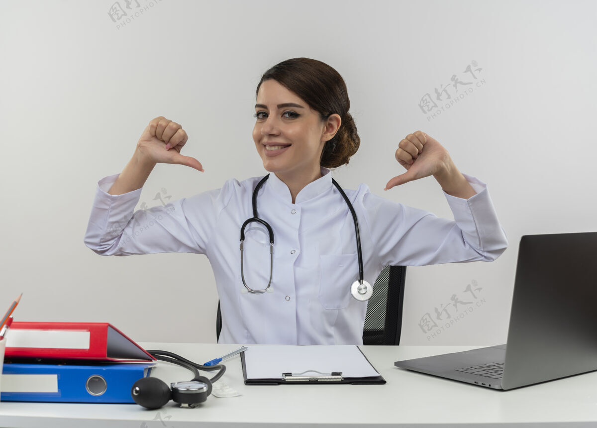 自己面带微笑的年轻女医生穿着医用长袍 手持听诊器坐在办公桌前 拿着医疗工具对着电脑指着自己的复印空间坐工具点