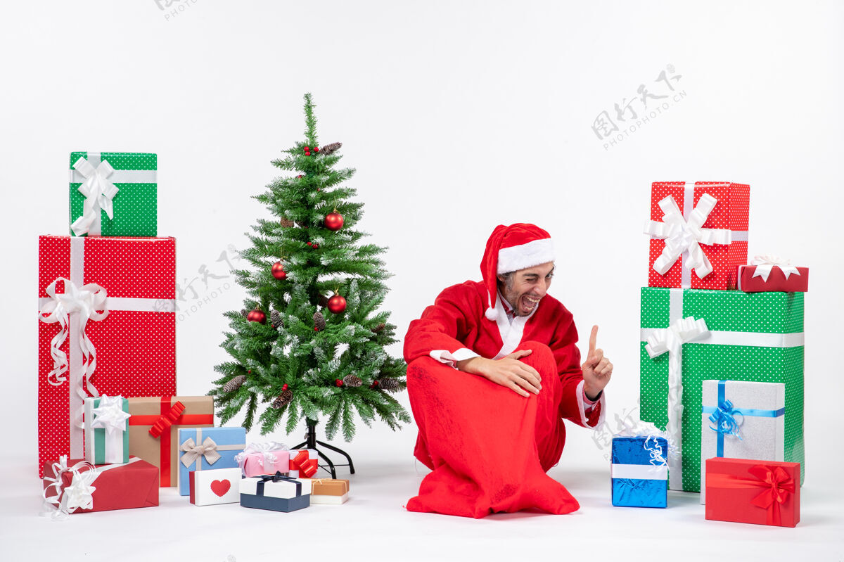 圣诞微笑有趣兴奋的年轻人打扮成圣诞老人与礼物和装饰圣诞树坐在地上指着上面的白色背景微笑男人礼物