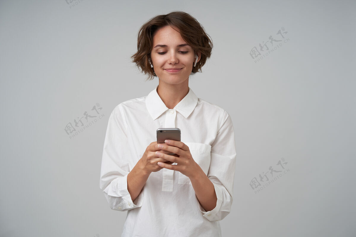 理发室内拍摄的一个可爱的年轻女子 棕色短发 穿着白衬衫 摆姿势 手里拿着智能手机 输入信息耳机表情肖像
