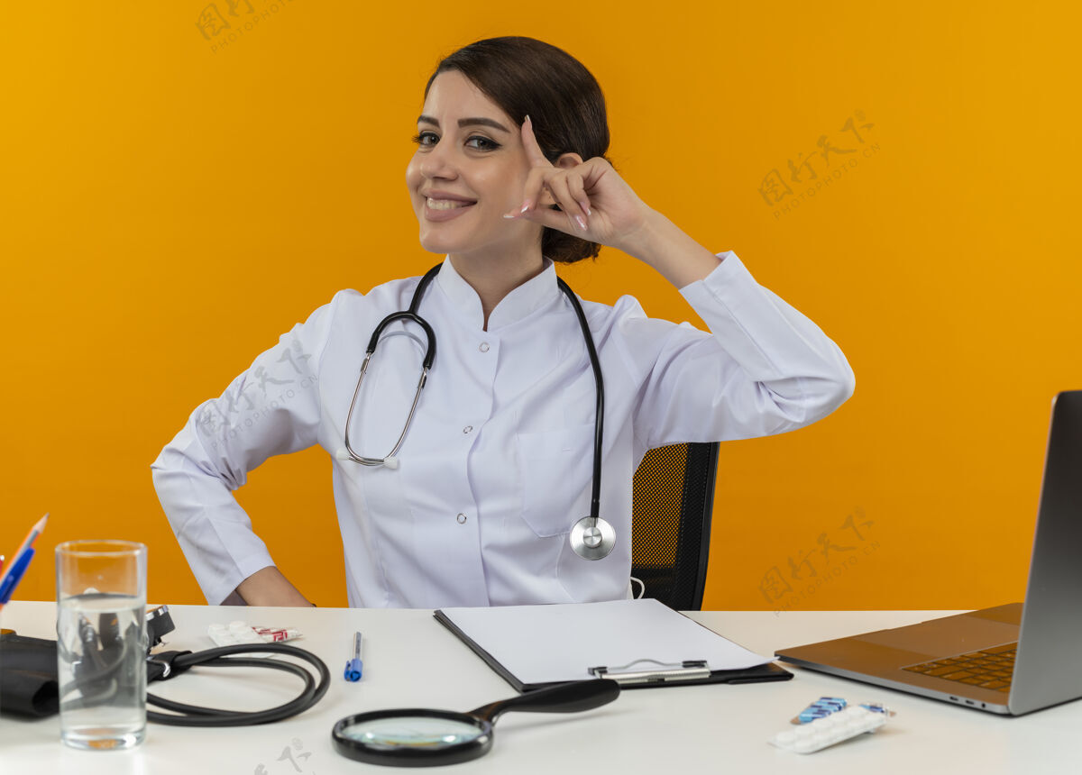 医生面带微笑的年轻女医生穿着医用长袍 手持听诊器坐在办公桌旁 手持医疗工具 将手指放在隔离黄色背景的额头上穿手指电脑