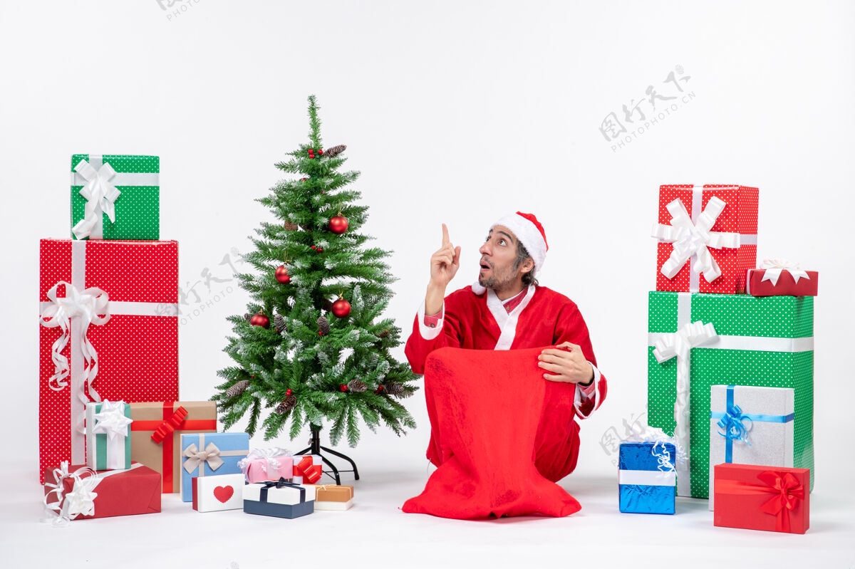 圣诞老人笑容满面的年轻人打扮成圣诞老人 拿着礼物和装饰好的圣诞树坐在地上 指着上面的白色背景人礼物圣诞老人