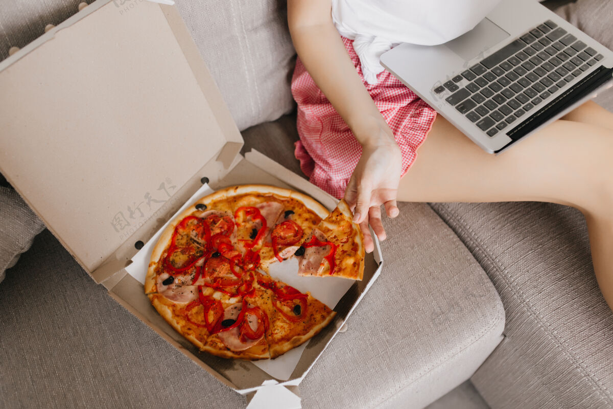 饥饿坐在沙发上拿着笔记本电脑和快餐的女士高加索女性自由职业者在工作时用电脑吃比萨饼厨房快速比萨饼