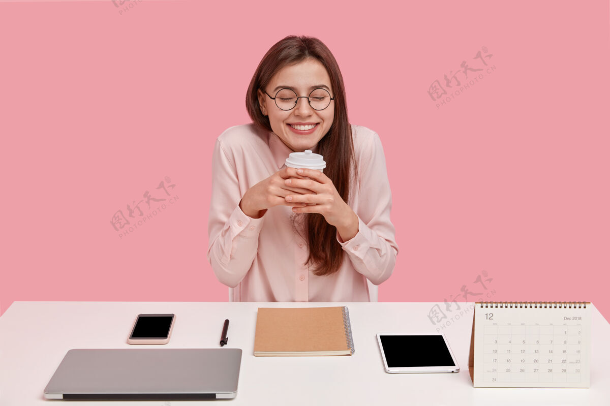 饮料喜出望外的年轻学生有着梦幻般的表情 拿着一次性咖啡杯 在同事的空间里休息 周围是现代化的笔记本电脑 平板电脑企业家专业人士高兴