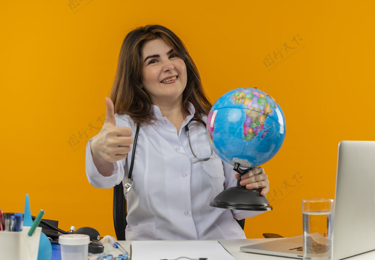 长袍微笑的中年女医生穿着医用长袍 手持听诊器 坐在办公桌旁 手持手提电脑 手持医疗工具 将大拇指放在孤立的橙色背景上 并留有复印空间拿着起来拇指